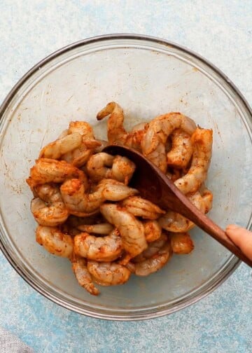a hand mixing raw shrimp with spices in a glass bowl.