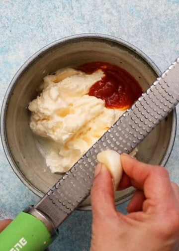 a hand grating one garlic clove over a bowl with white mayo and red sriracha sauce.