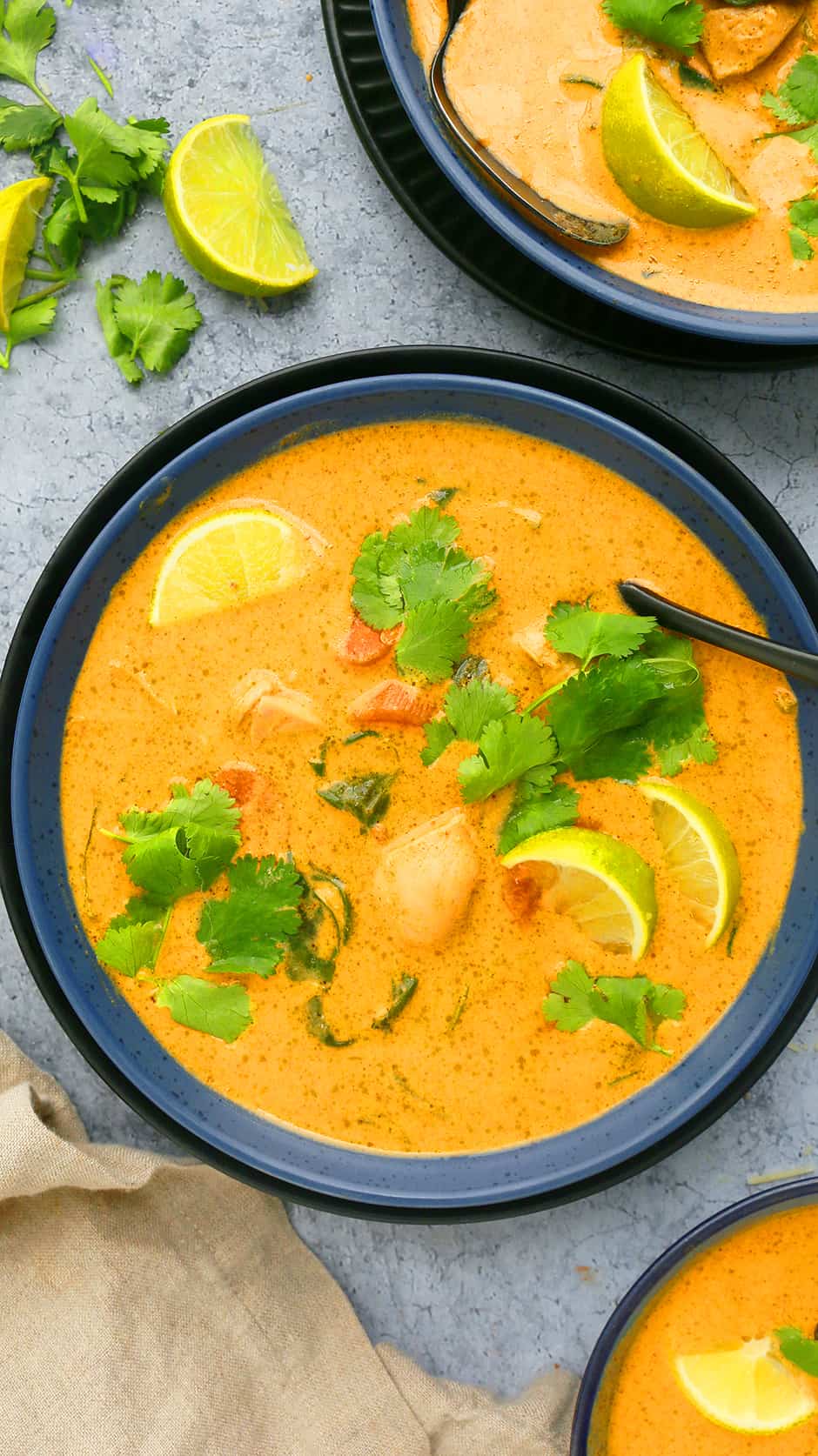 a blue bowl with red curry chicken soup garnished with lime wedges and cilantro
