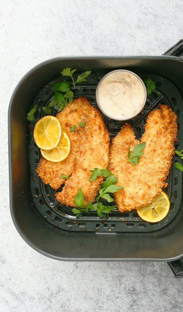air fryer basket with tilapia with a side of sauce in a small bowl and garnished with parsley and lemons