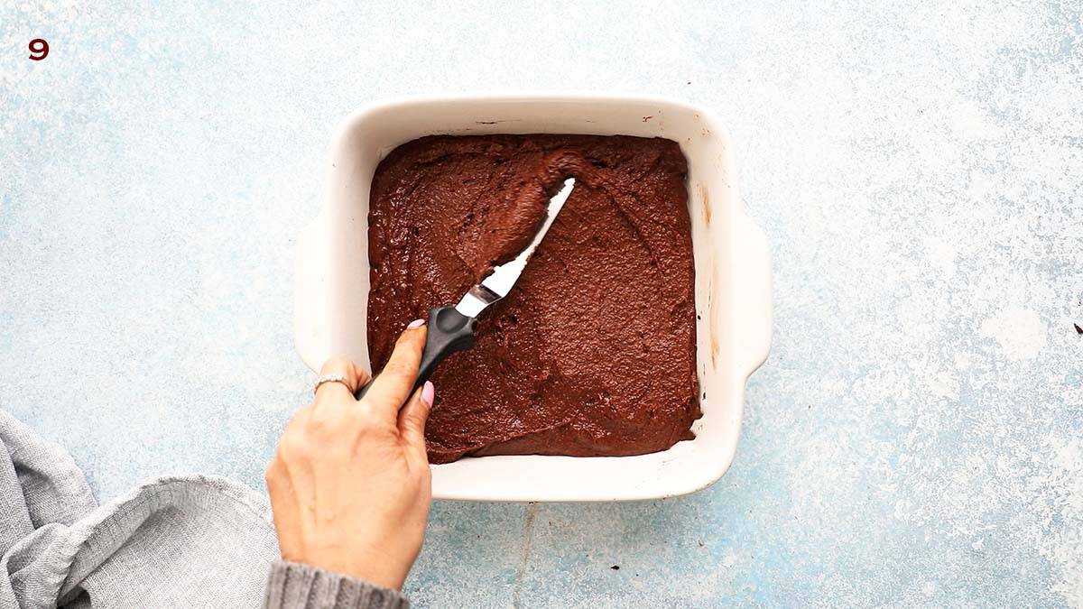 a hand spreading brownie batter in a white square baking dish.