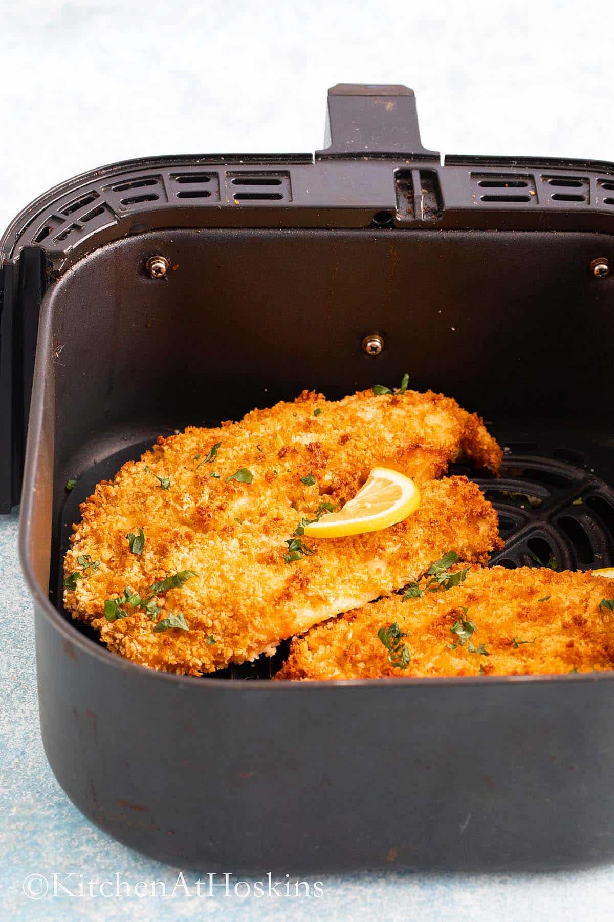 breaded tilapia fillets in an air fryer basket topped with lemon slice.