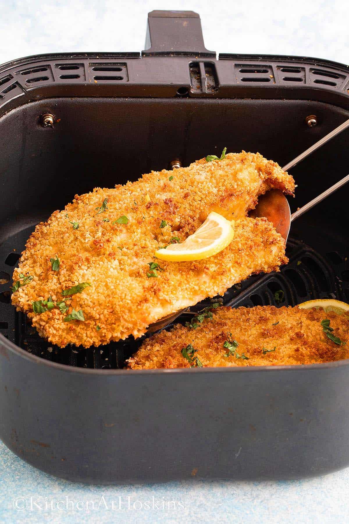 Removing a breaded fish fillet from an air fryer basket using a flat spatula.