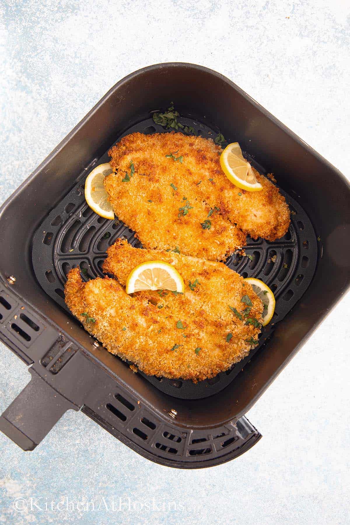 2 breaded fish fillets in an air fryer basket garnished with lemon slices.