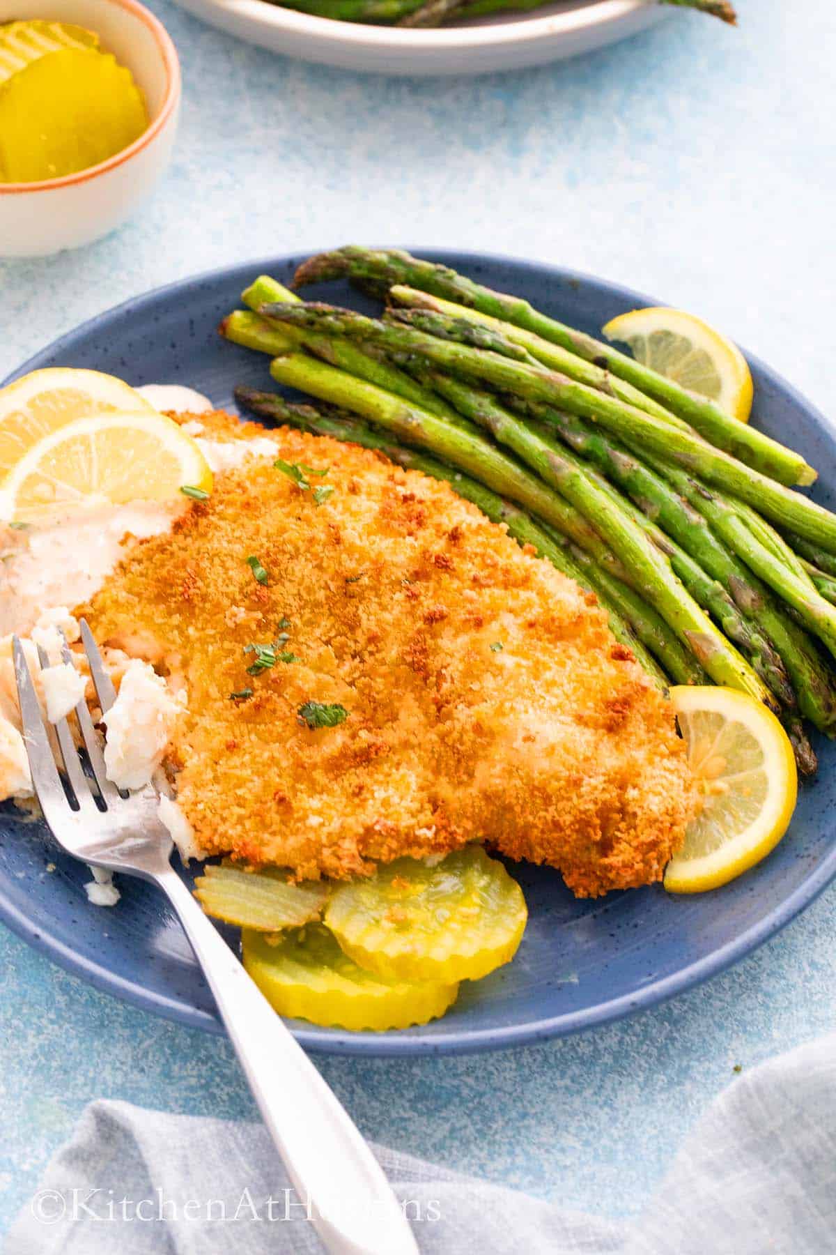 blue plate with cooked fish, aspargus, pickles and a fork.