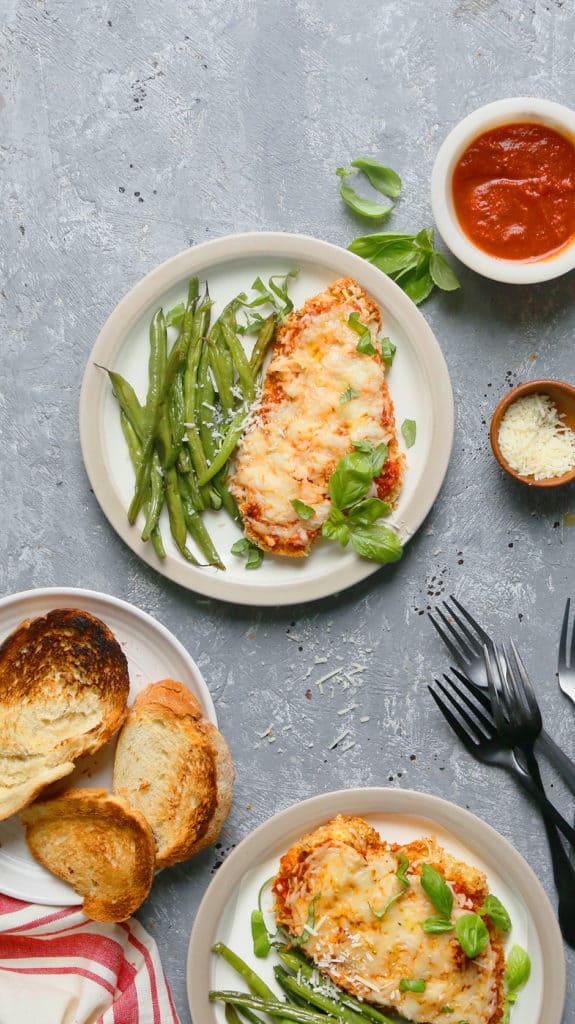 white plates with air fryer chicken parmesan along with green beans, forks, toasted bread, fresh basil and marinara sauce