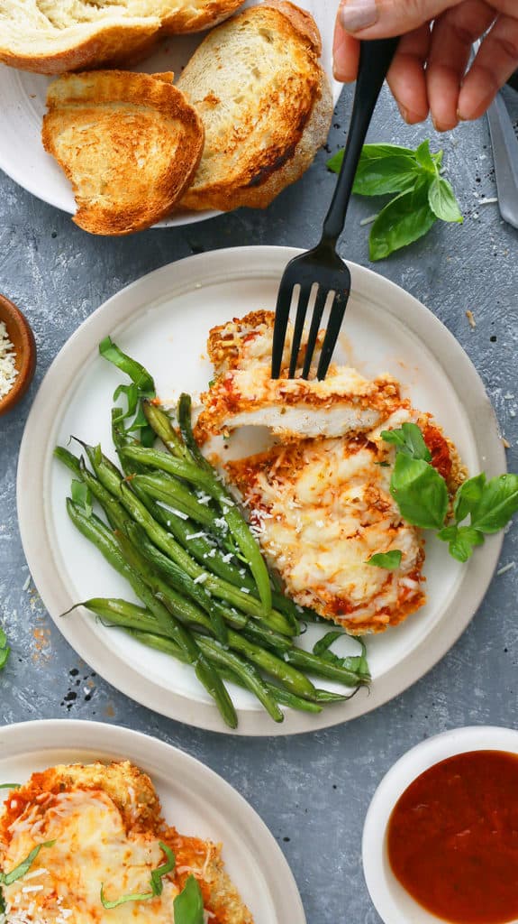 a black fork trying to take a bite off chicken parm in air fryer along with green beans, forks, toasted bread, fresh basil and marinara sauce