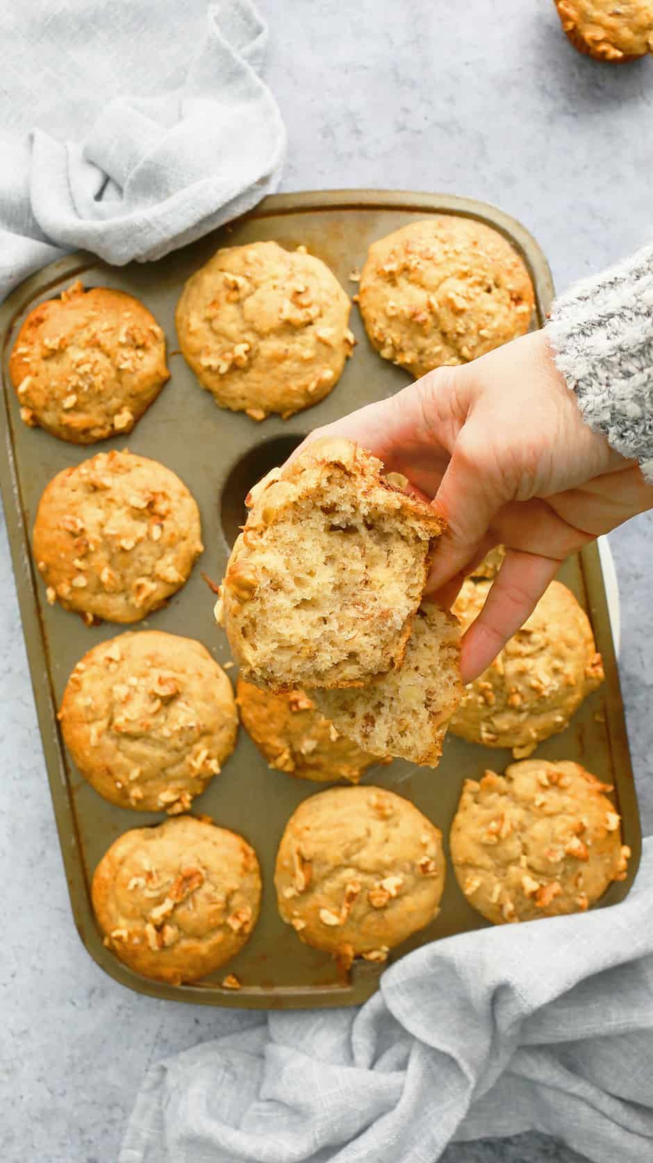 a hand holding half a banana muffin above a pan full of muffins