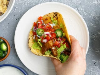 a hand grabbing a taco filled with spicy potatoes from a white plate