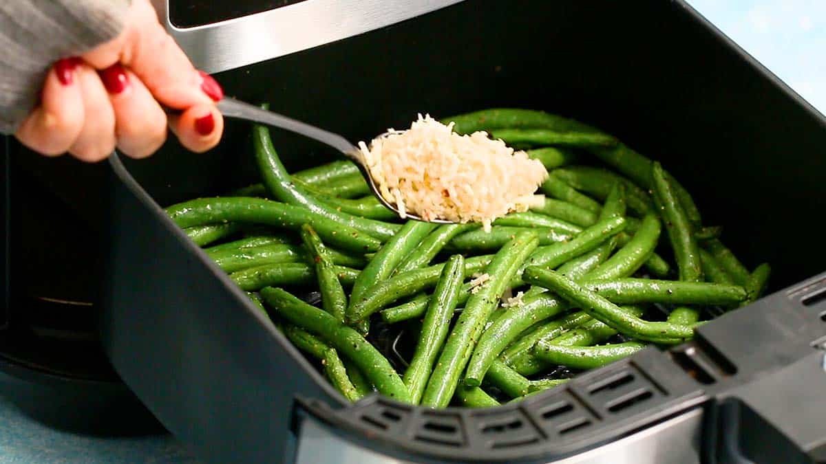 a hand sprinkling parmesan cheese on top of green beans in air fryer basket.