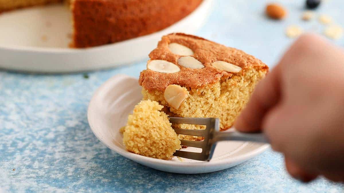 a hand cutting a piece of cake from a small white plate with a fork.