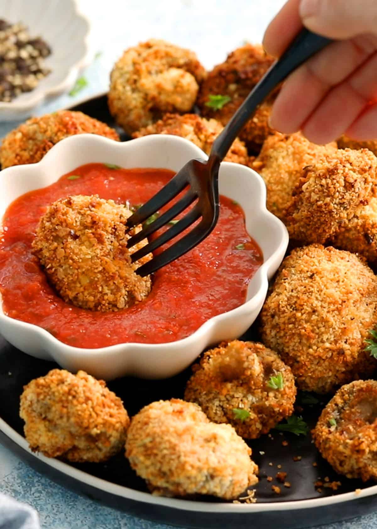 a hand dipping one breaded mushroom in red sauce placed in a white bowl. 