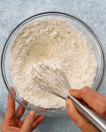 two hands whisking white flour in a large glass bowl.