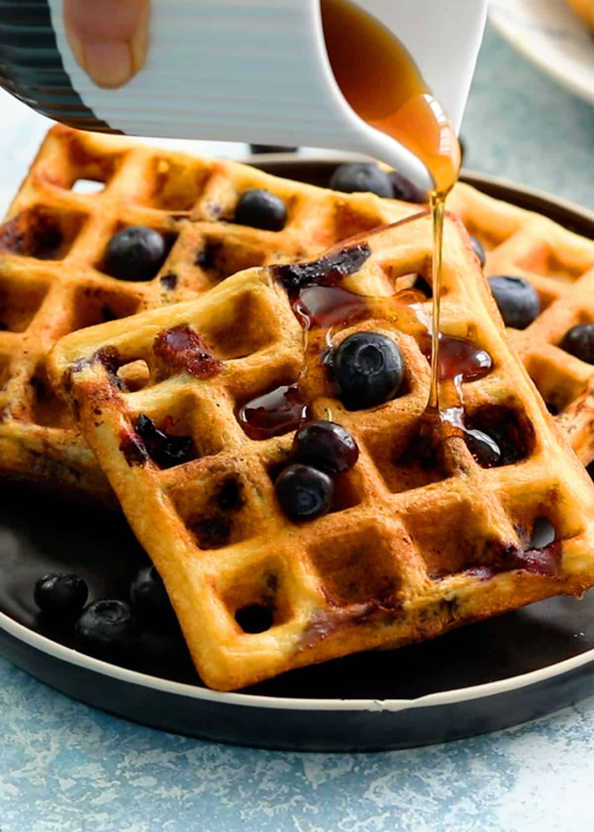 a hand pouring golden syrup on top of 3 waffles placed on a black plate.