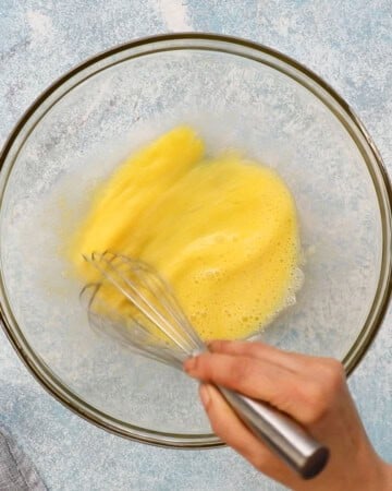 a hand whisking yellow eggs in a glass bowl.