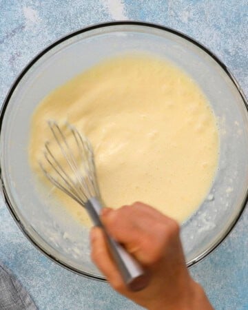 a hand whisking white liquid in a large glass bowl.