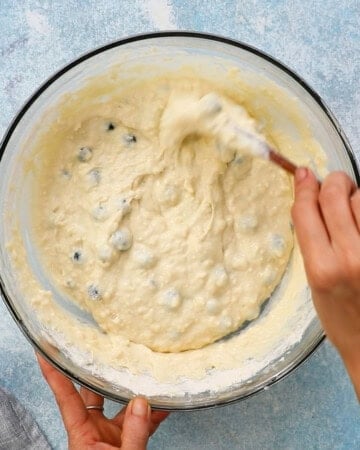 two hands mixing blueberry waflle batter in a large glass bowl.