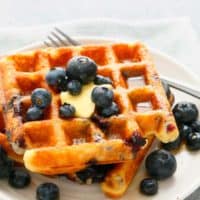 homemade blueberry waffles on a white plate topped with butter, maple syrup and more blueberries