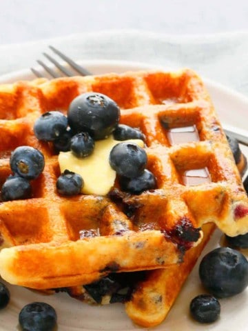 homemade blueberry waffles on a white plate topped with butter, maple syrup and more blueberries