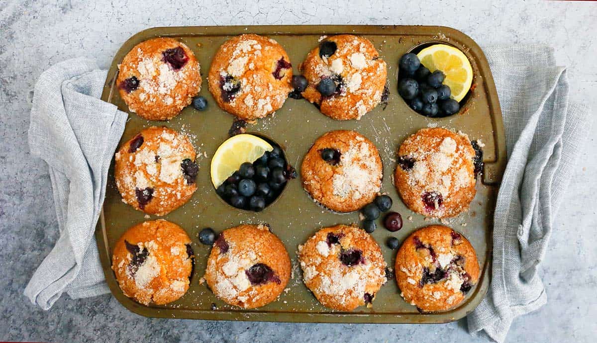 a muffin pan with baked lemon blueberry muffins along with folded grey kitchen towels on both sides