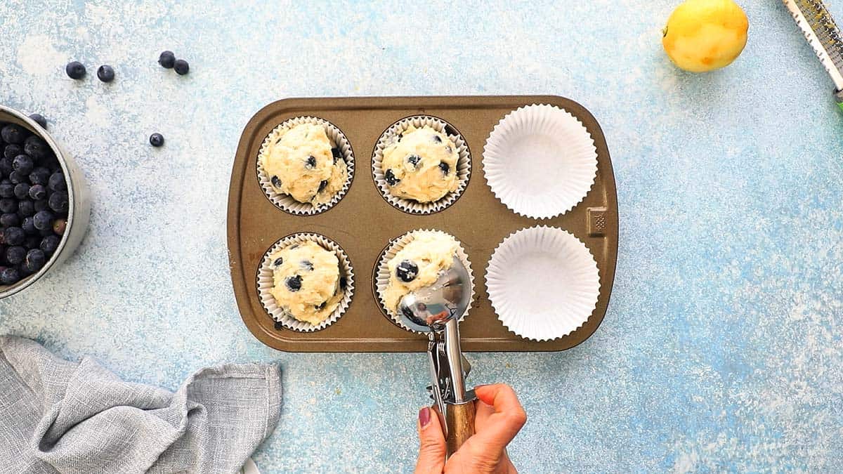 one hand filling a 6 cup muffin pan with batter using a stainless steel ice cream scoop.