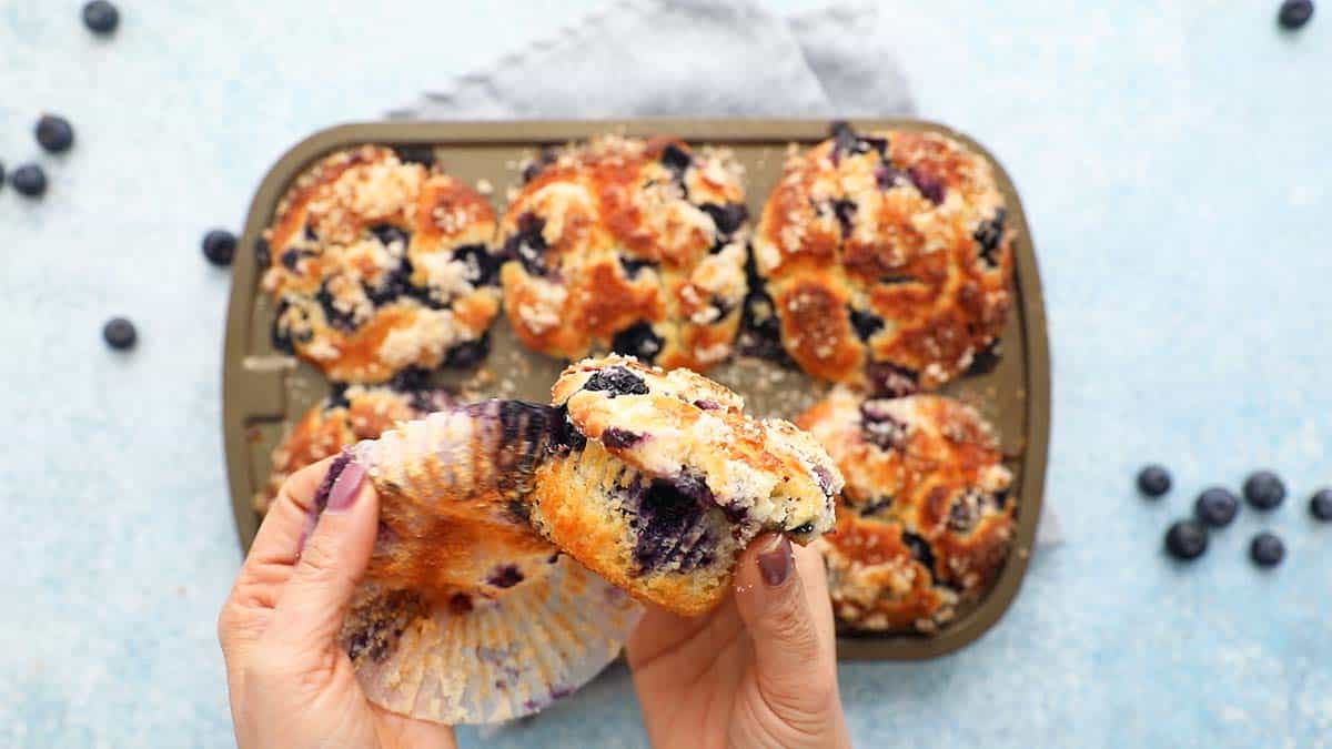 two hands peeling off the paper liner from a blueberry muffin.