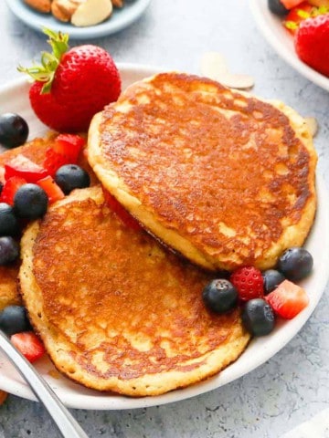 white plate with 3 almond pancakes along with a fork and fresh berries