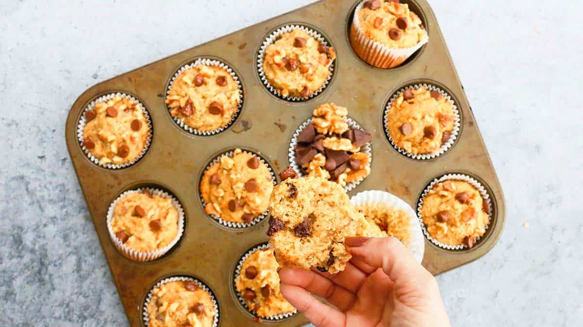 a hand holding a split banana almond muffin above a muffin pan filled with the same. 