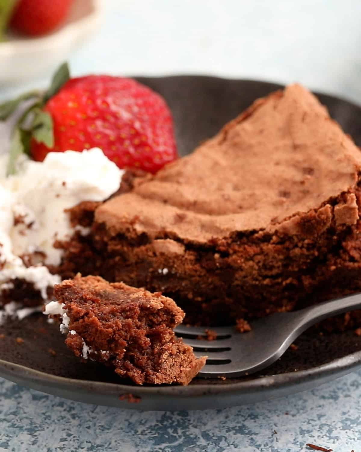 one slice of chocolate cake in a black plate along with one strawberry and whipped cream.