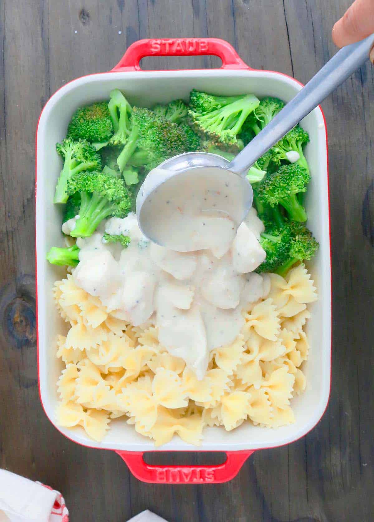 pouring chicken alfredo sauce on a baking dish with cooked pasta and broccoli 