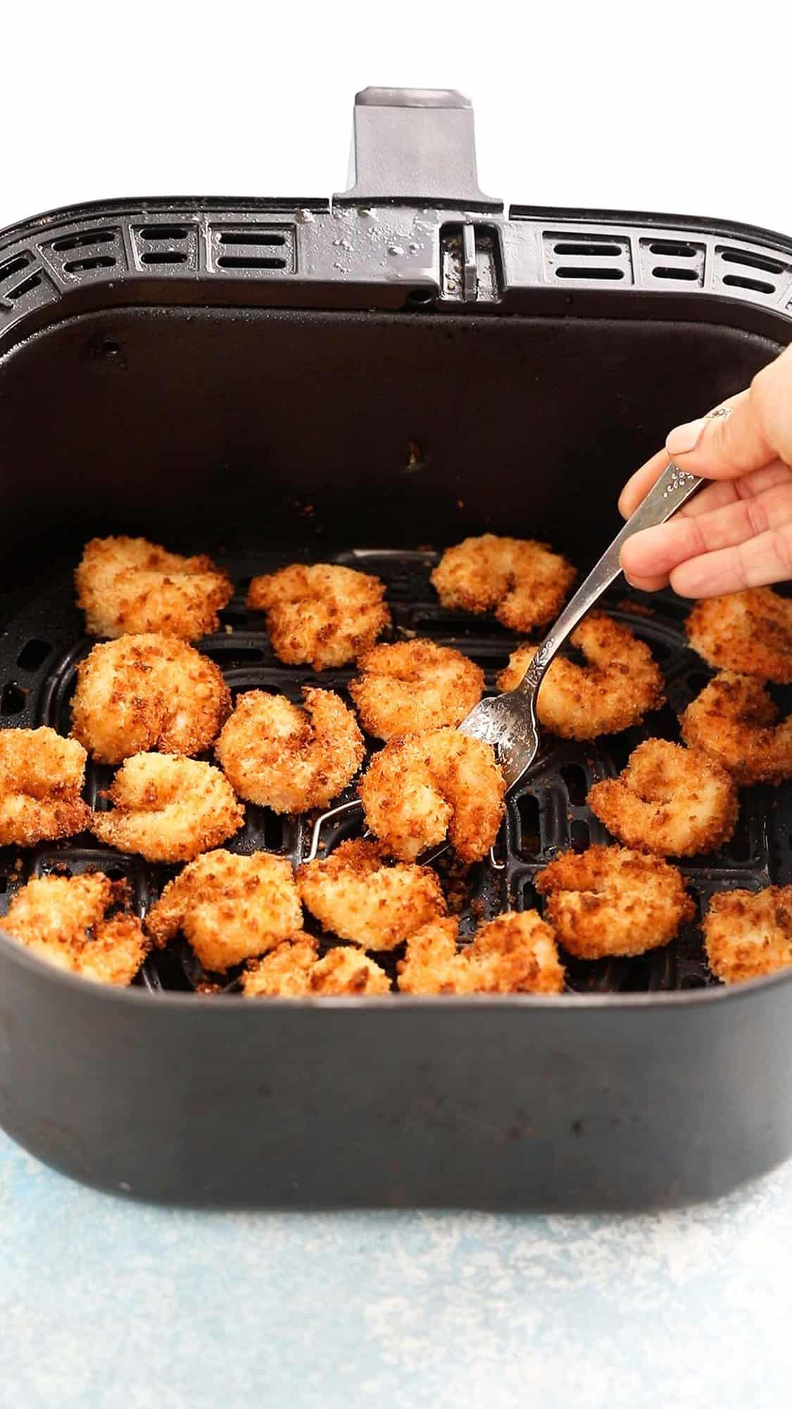 a hand lifting one breaded shrimp from an air fryer basket using a fork.