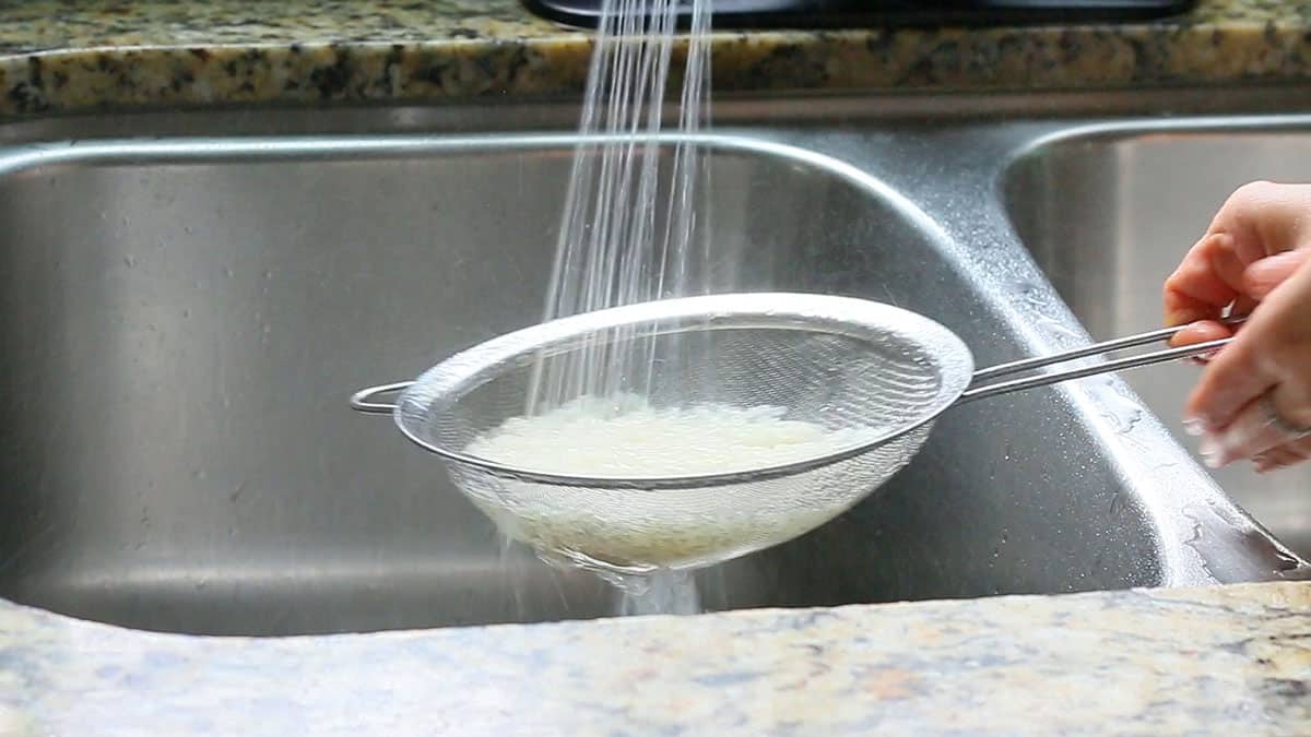 raw white rice in a metal strainer placed under running tap water.