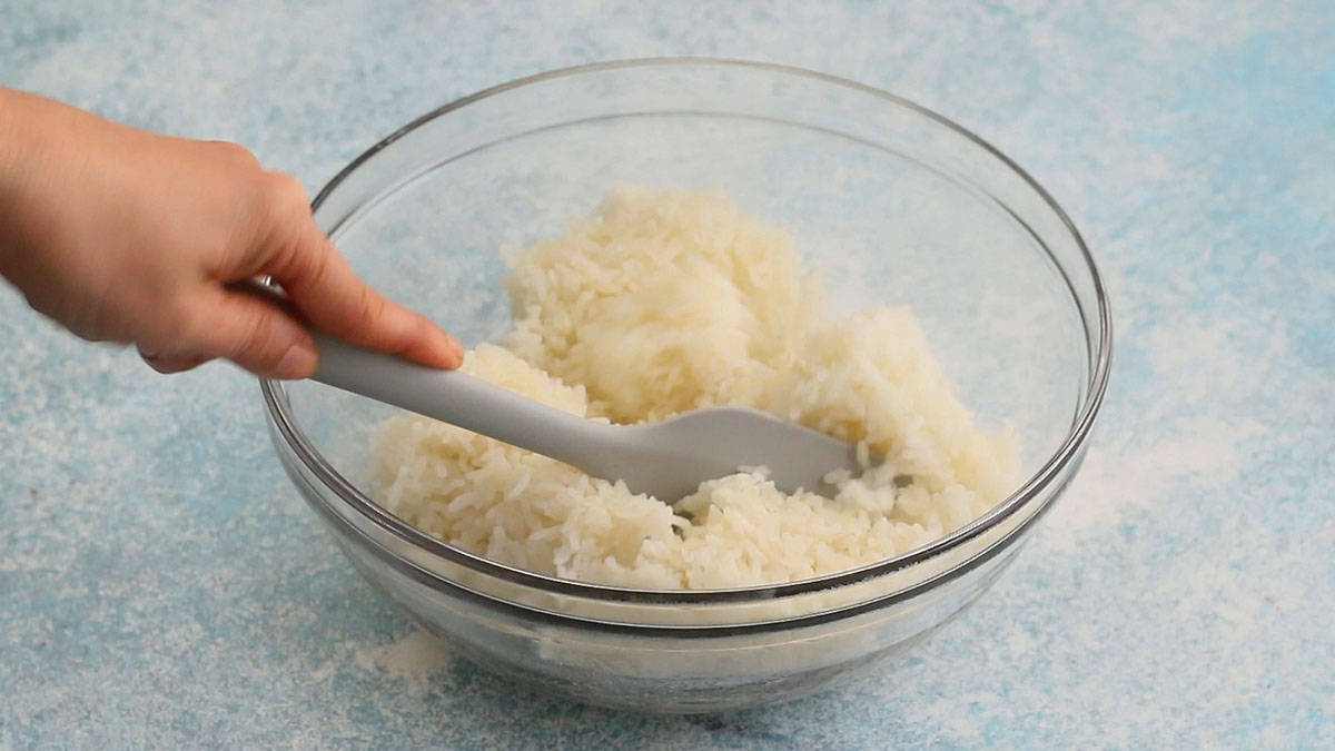 a hand mixing cooked white sushi rice in a glass bowl using a grey spatula.