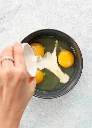 a hand pouring milk into a blue bowl with 3 eggs.