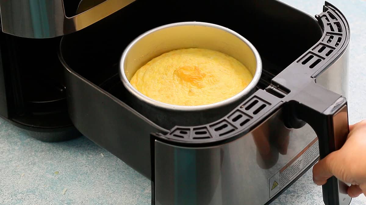 a hand holding the hand of an air fryer basket with a metal pan with omelette.