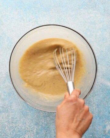 a hand whisking banana cake batter in a glass bowl.
