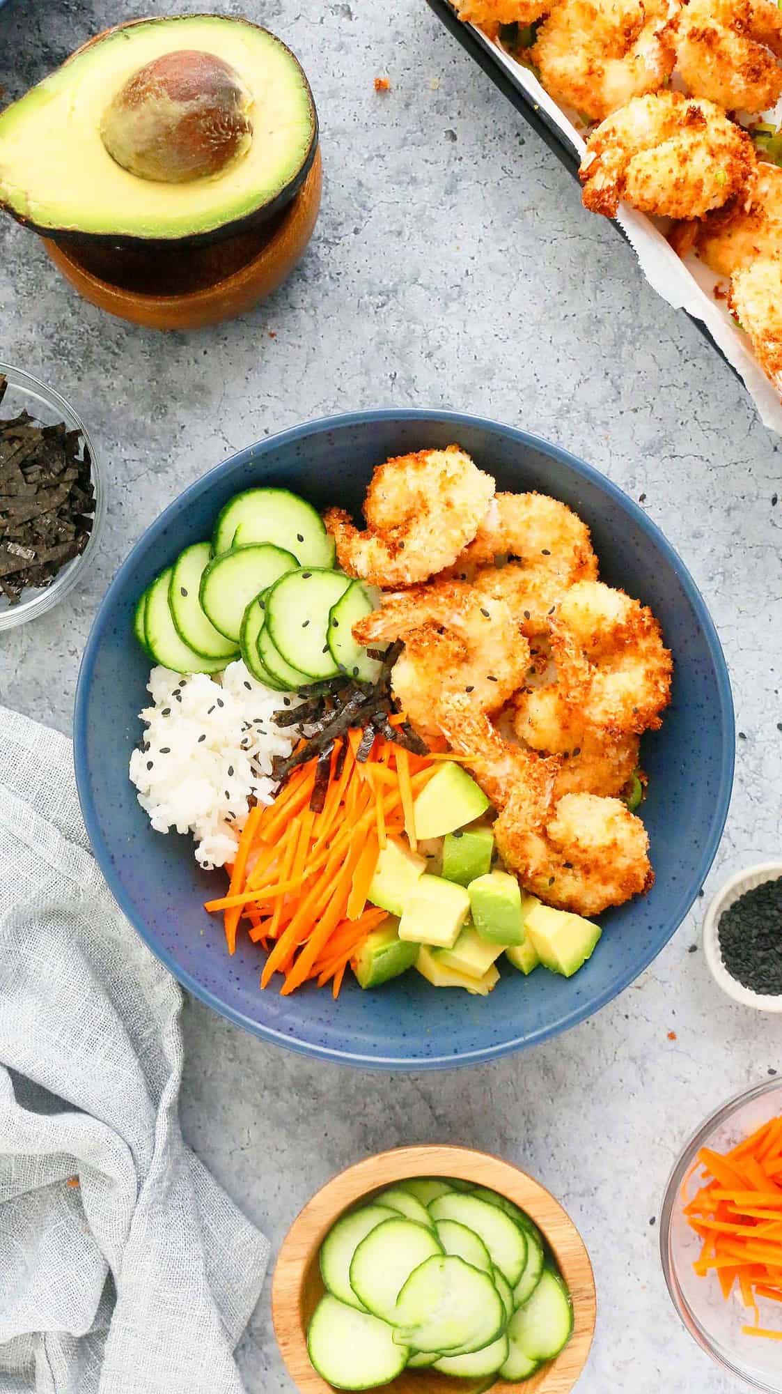 blue bowl with cooked shrimp, sliced cucumber, julienned carrot, chopped avocado and sushi rice. 