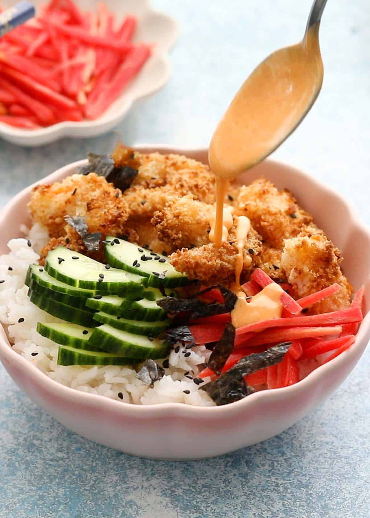a hand drizzling pink sauce over a pink bowl layered with shrimp, cucumber, rice and carrot.