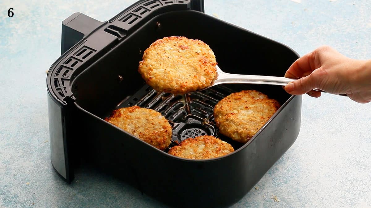 a hand holding one cooked chicken burger patty with a flat spatula above an air fryer basket.