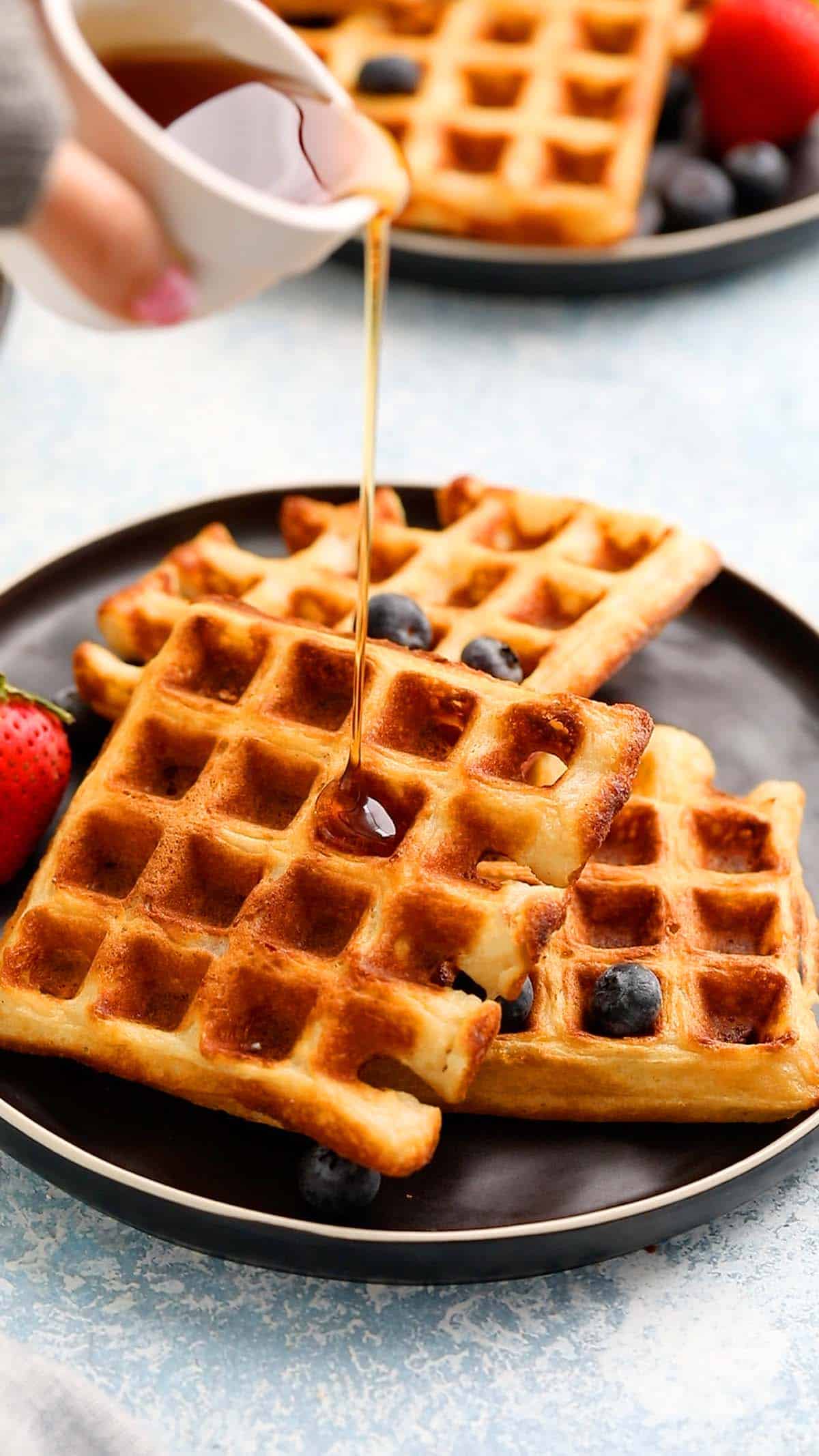 a hand pouring maple syrup over cooked waffles placed on a black plate.