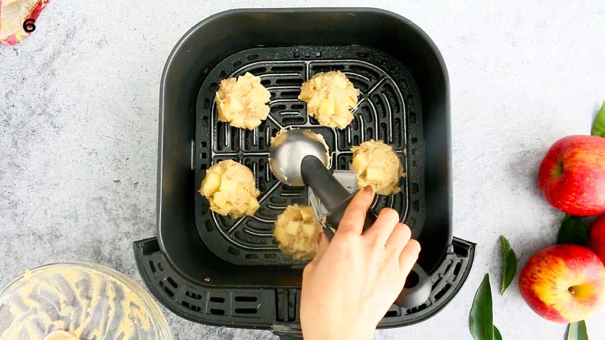 one hand adding fritter dough into an air fryer basket using an ice cream scoop.