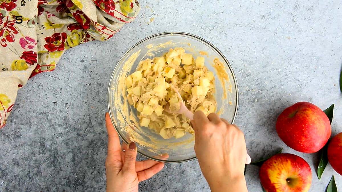 two hand stirring chopped apples into a batter using a pink spatula.