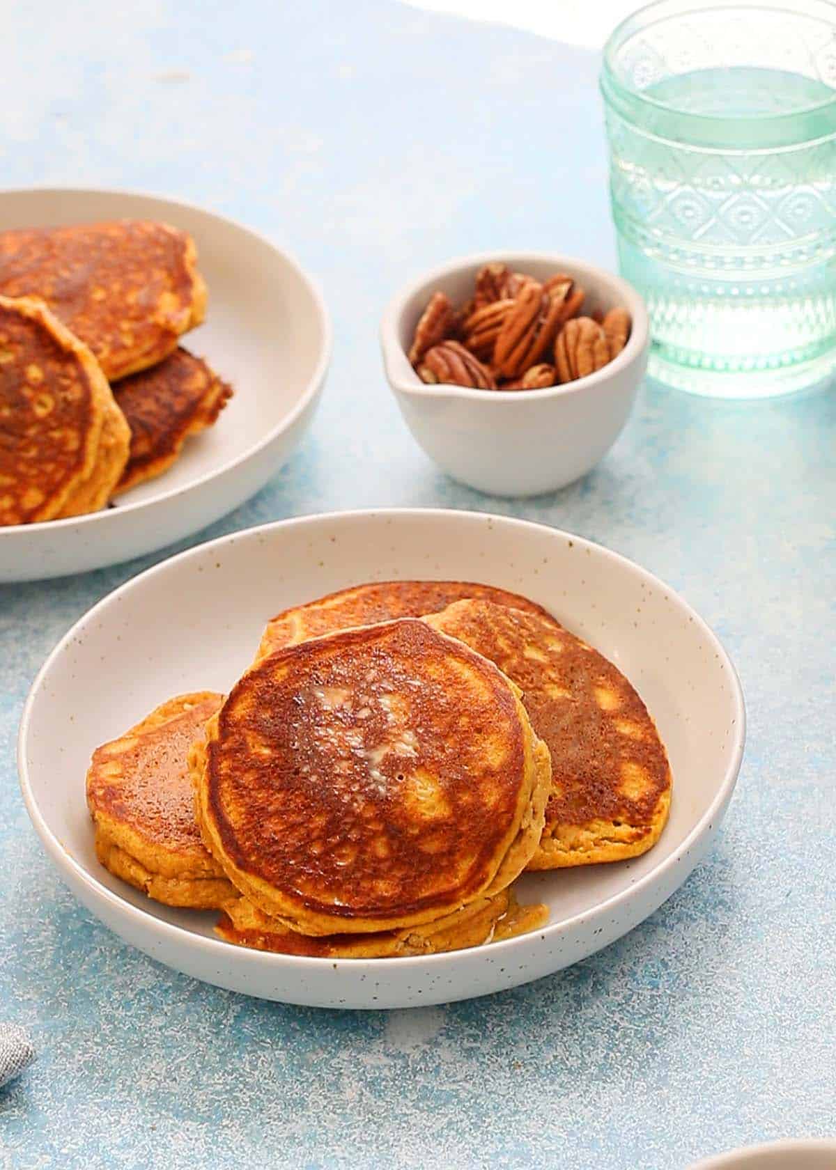 3 pancakes in a white plate along with   a bowl of pecans and a glass. 