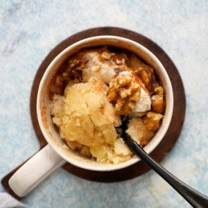 cooked apple cake in a white mug along with a black spoon.