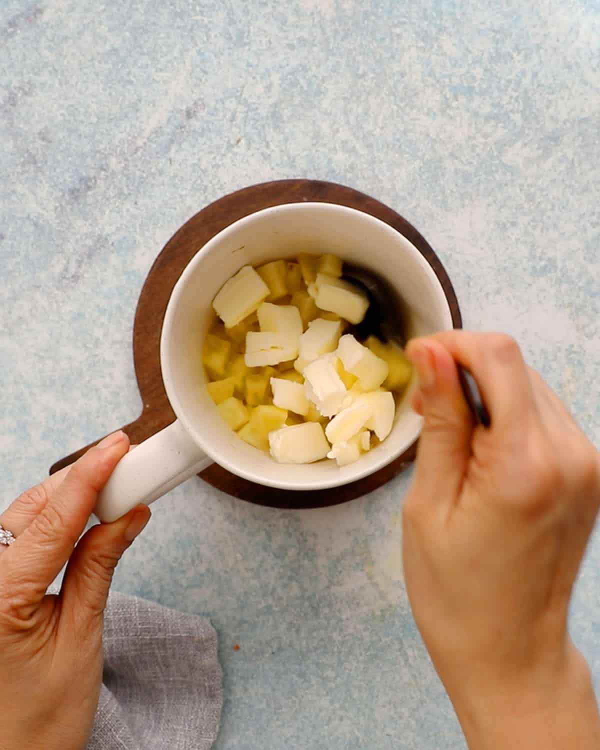 two hands mixing chopped apple with butter in a white mug.