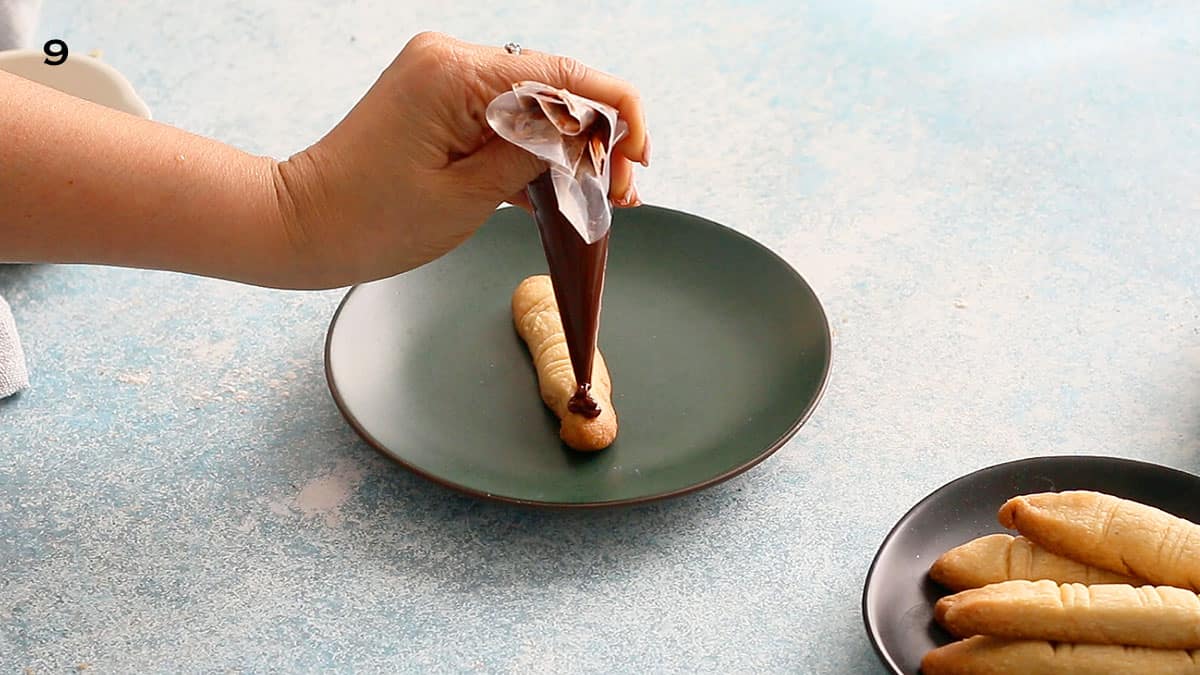 a hand squeezing melted chocolate on top a finger shaped cookie.