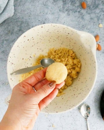 a hand holding one cookie dough above a white bowl.