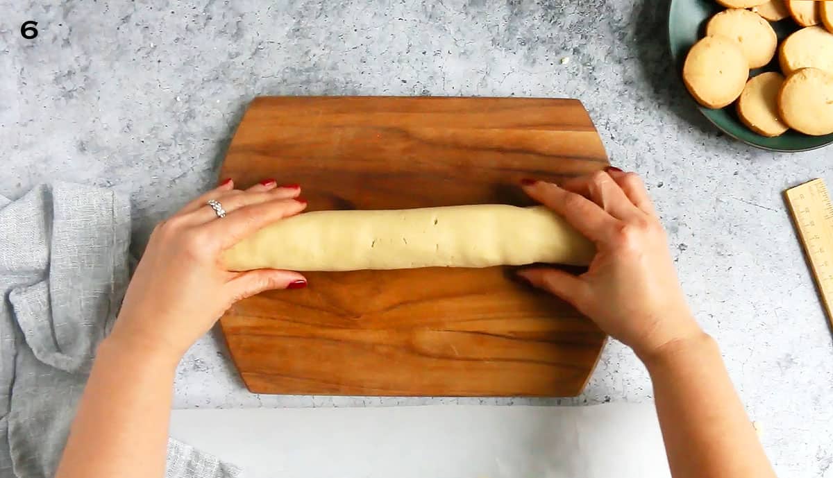 2 hands shaping cookie dough into a log on a wooden board.