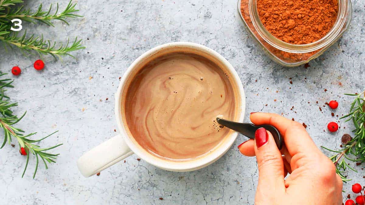 a hand stirring homemade mocha in a white mug using a spoon.