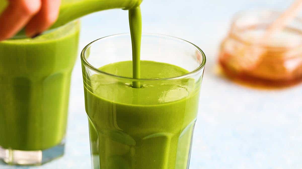 A hand pouring green spinach smoothie from a bottle into a glass.